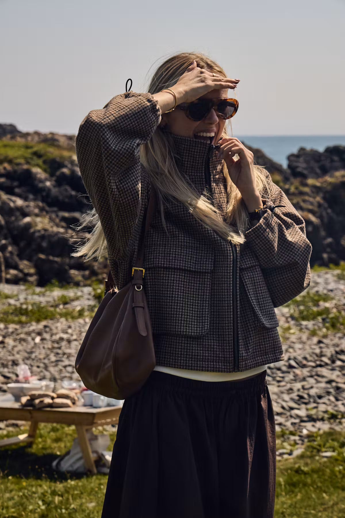 ICHI Esolu Jacket in Brown Check - a woman standing on a rocky outdoor beach or cliffside wearing a dark brown and black checkered zip-up jacket, a long dark skirt, and sunglasses, with a brown shoulder bag.