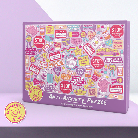 A purple rectangular jigsaw puzzle box labeled 'ANTI-Anxiety PUZZLE' with a dense collage of pastel and bright stickers, signs, and therapy-themed illustrations on the front. A round yellow sticker badge reading 'ANTI-Anxiety SOCIETY' appears in the lower left on a lavender background.