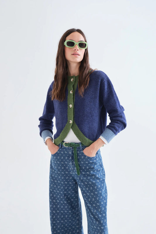 A woman modeling a navy blue knit cardigan with green trim and buttons, paired with blue patterned pants, a white top, and green sunglasses against a plain white studio background.