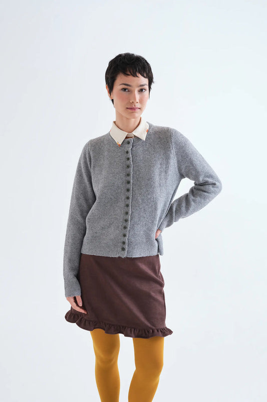 A model wearing a gray button-front cardigan over a cream collared shirt, paired with a brown ruffled mini skirt and mustard yellow tights, against a plain white studio background.