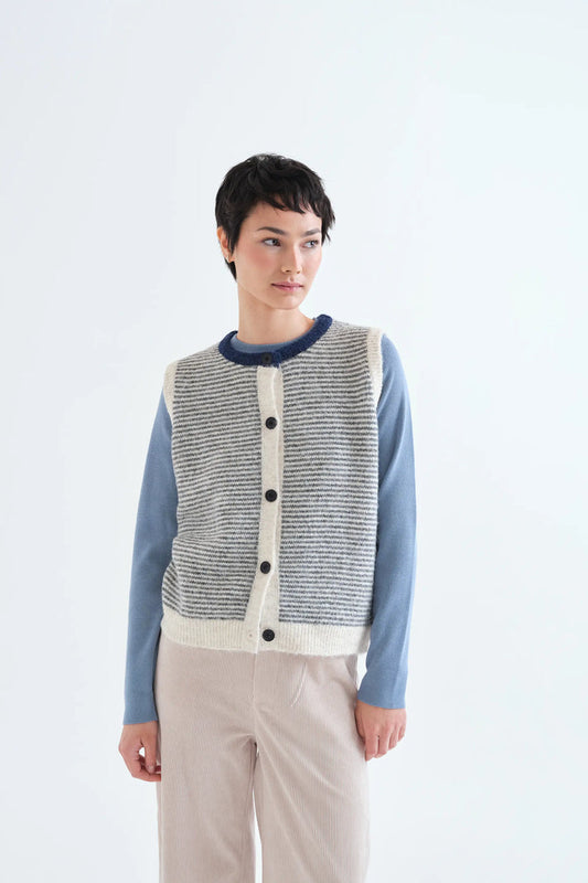 A model wearing a gray and white textured knit cardigan vest over a dusty blue long-sleeve shirt and beige trousers, set against a plain white studio background.