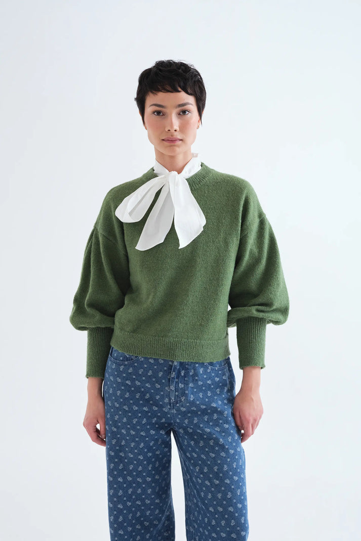 A model wearing an olive green knit sweater with a large white bow at the collar, paired with blue patterned pants, against a plain white studio background.