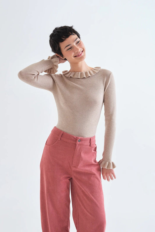 A model wearing a beige long-sleeve knit top with a ruffled neckline and cuffs, paired with dusty rose wide-leg trousers, against a plain white studio background.