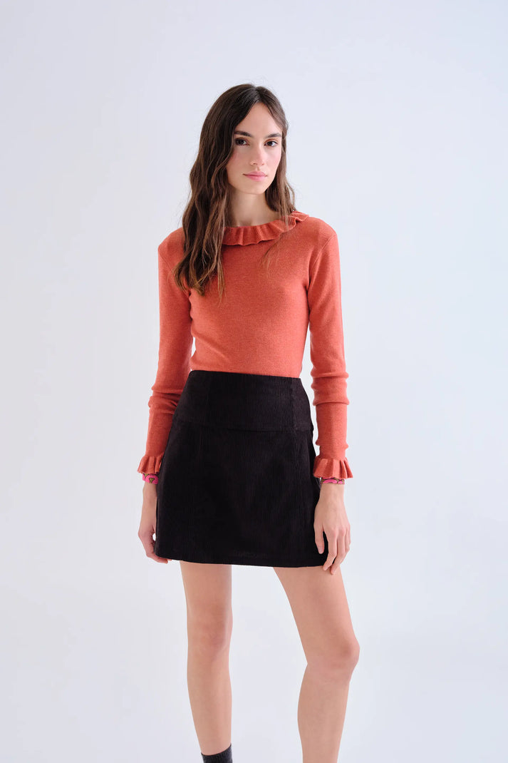 A woman modeling a long-sleeve knit top in a muted orange-coral color with ruffled trim at the neckline and cuffs, paired with a black mini skirt, against a plain light gray studio background.