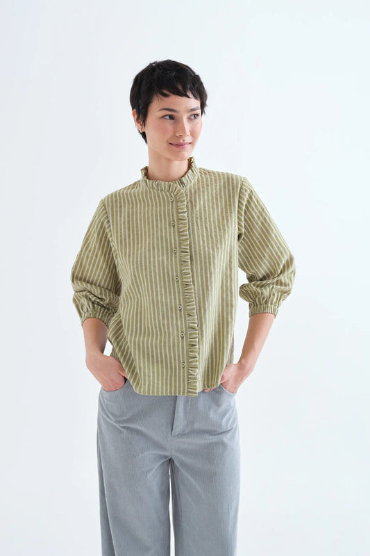 A woman modeling a light olive green and white vertically striped blouse with ruffle trim and gathered sleeves, paired with light gray trousers, against a plain white studio background.