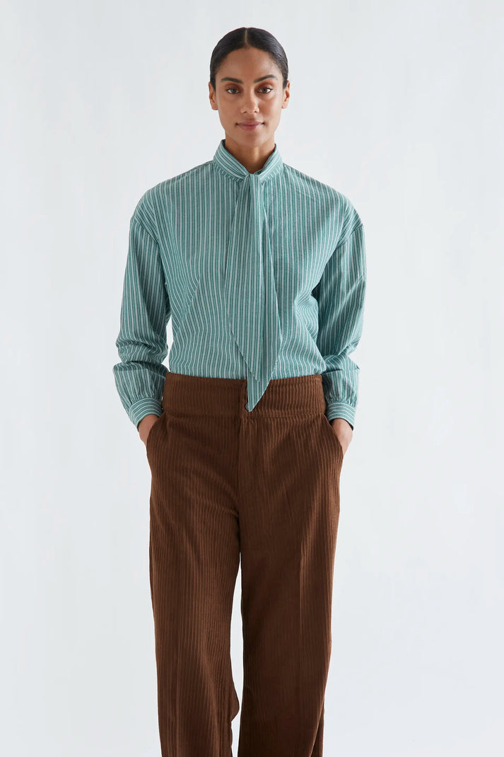 A woman modeling a light blue-green and white vertically striped button-down shirt with a high collar, paired with brown corduroy trousers, against a plain white studio background.