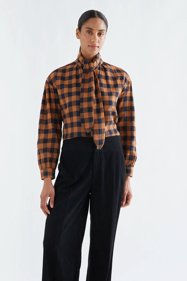 A model wearing a brown and black checkered long-sleeve blouse with a high neck and front tie detail, paired with black pleated trousers, against a plain white studio background.