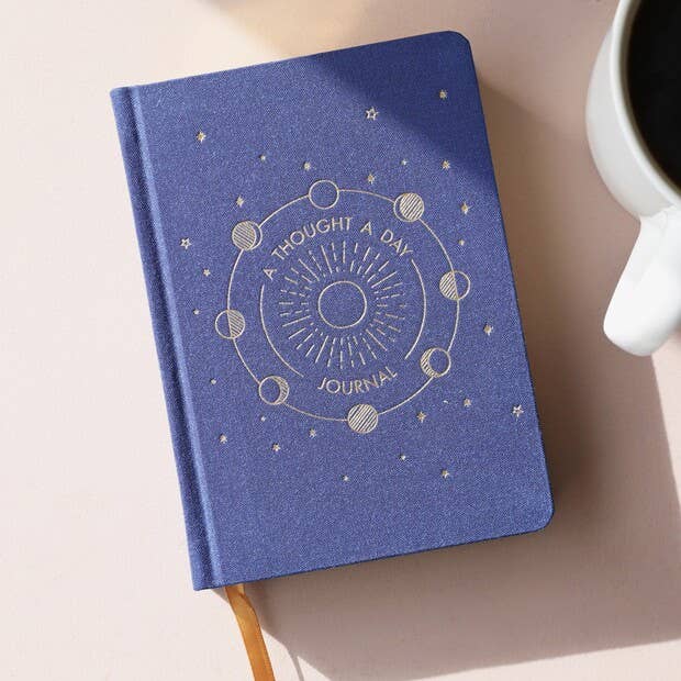 Blue journal with celestial design on a surface next to a white mug.