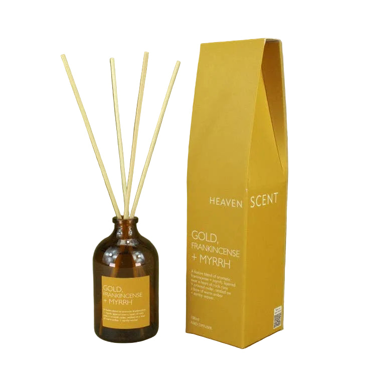 An amber glass reed diffuser bottle with four natural reed sticks stands next to its mustard yellow packaging box on a solid olive green background. The box is branded 'HEAVEN SCENT' and labeled 'GOLD FRANKINCENSE + MYRRH'.