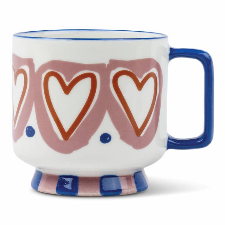 A white ceramic mug with a blue handle and rim, decorated with a pink pattern of interlocking heart outlines and blue dots, and a blue and pink striped footed base.