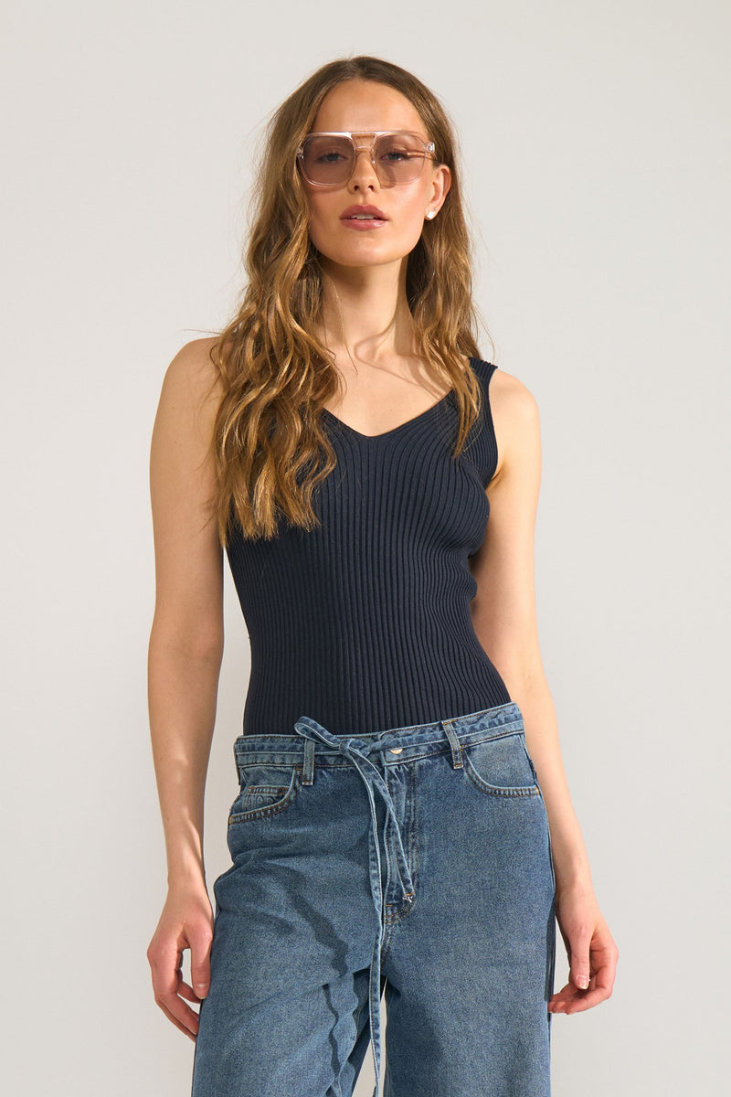 Woman wearing a navy ribbed tank top and blue denim pants against a plain background