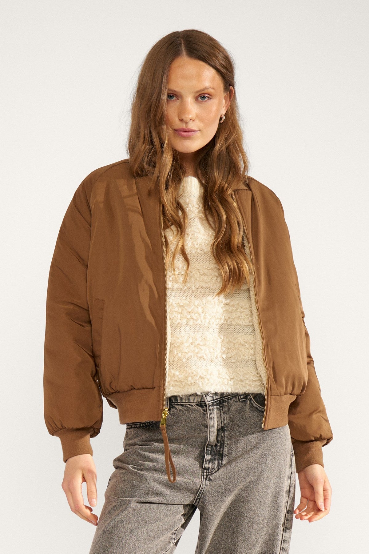 Numph Etera Reversable Jacket in Dachshund - a woman wearing a brown bomber jacket over a cream textured sweater and grey jeans, standing against a plain light background.