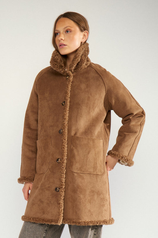 A woman wearing a brown suede-like shearling coat with a high fuzzy collar, front buttons, and large patch pockets, photographed against a plain light gray studio background.