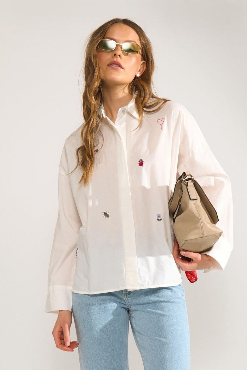Woman wearing a white shirt with embroidered designs, light blue jeans, and holding a beige handbag against a white background.