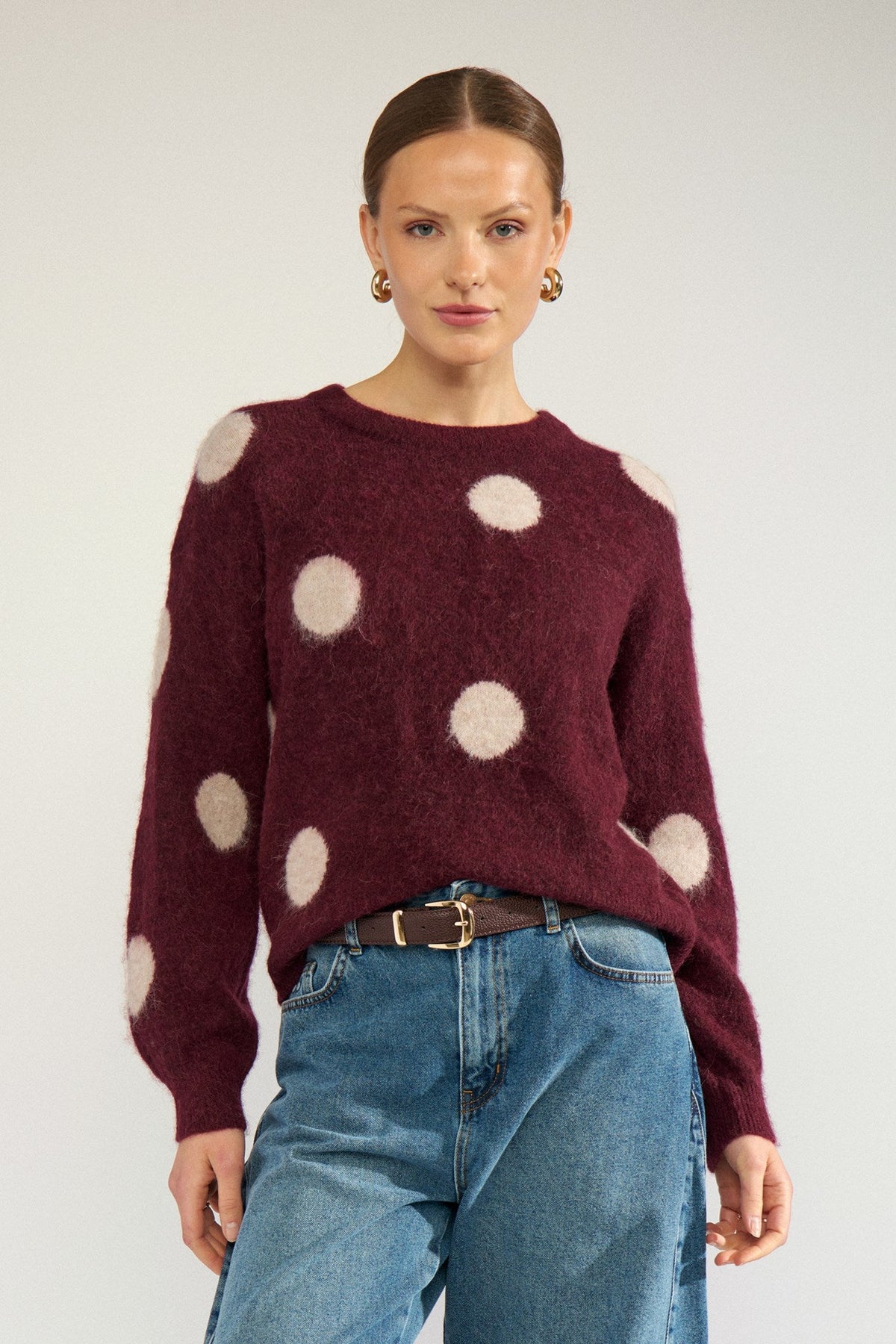 Numph Suri Dot Pullover in Fig - a woman modelling a fuzzy long-sleeve sweater in deep burgundy with large cream polka dots, paired with blue jeans and a brown belt, against a light grey studio background.