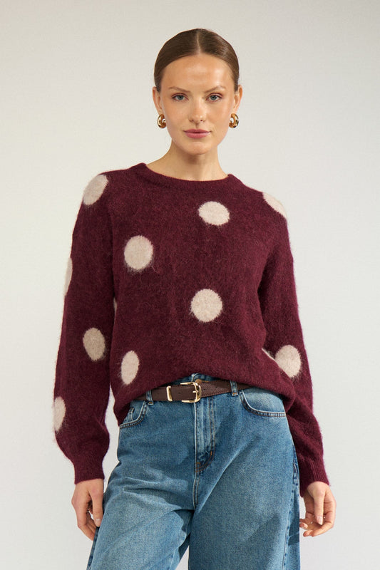 Numph Suri Dot Pullover in Fig - a woman modelling a fuzzy long-sleeve sweater in deep burgundy with large cream polka dots, paired with blue jeans and a brown belt, against a light grey studio background.