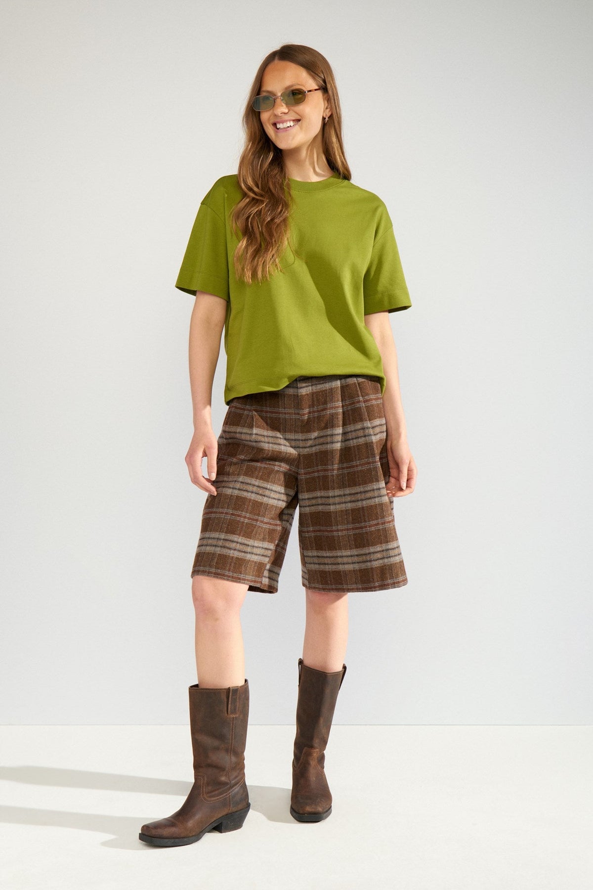 Numph Ezma Shorts in Kelp Check - a woman modelling an olive green short-sleeve t-shirt, brown plaid knee-length shorts, and dark brown leather knee-high boots against a plain light grey studio background.