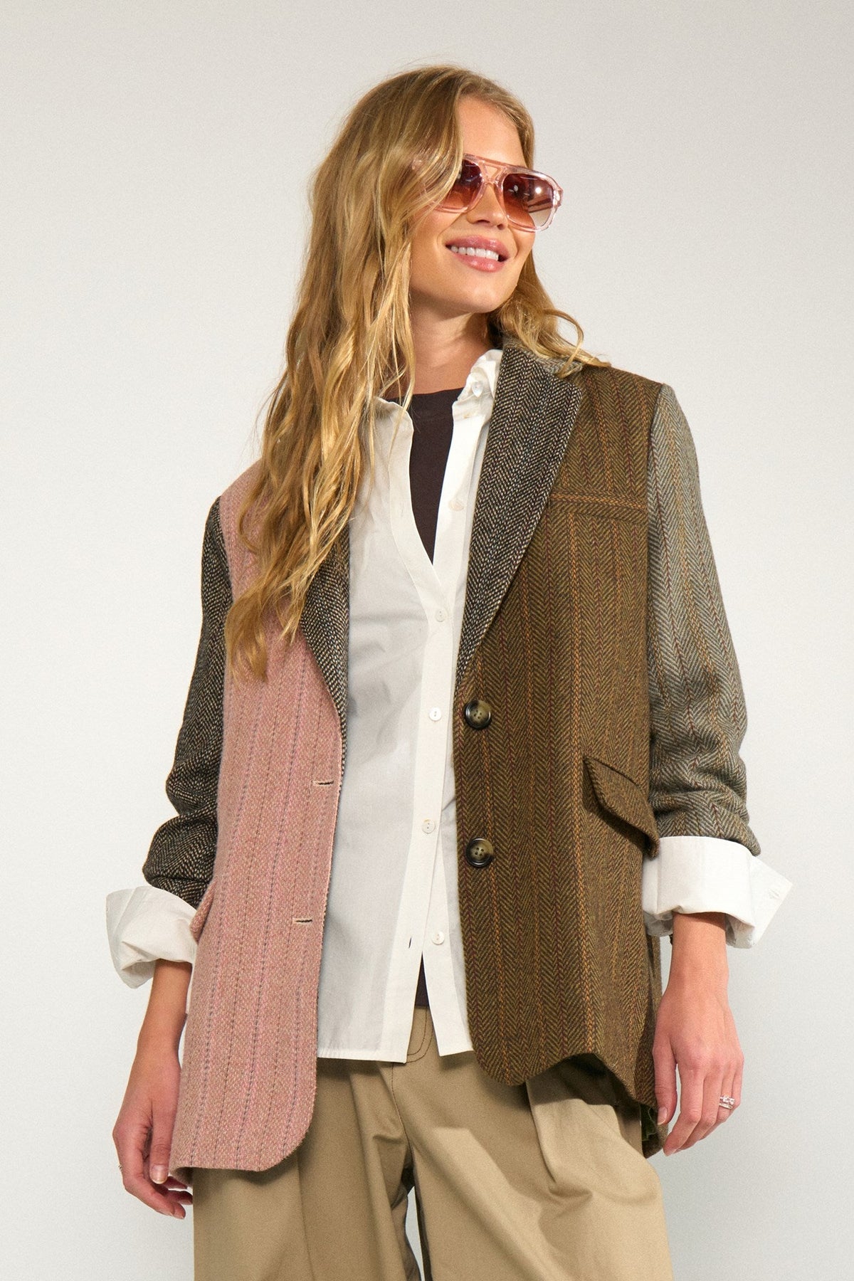 A woman wearing a multi-textured patchwork blazer vest in brown, pink, and gray, layered over a white button-down shirt and beige trousers, with pink-tinted sunglasses, against a plain light gray background.