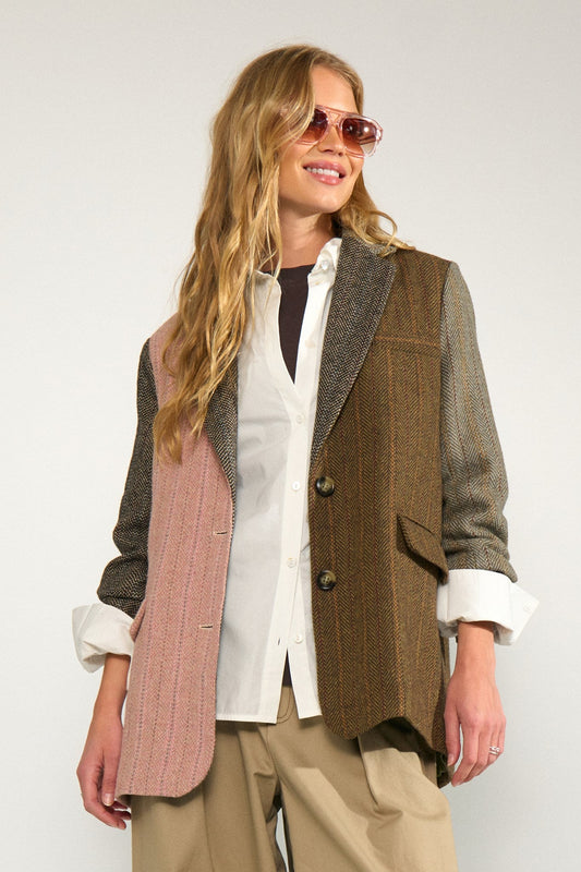 A woman wearing a multi-textured patchwork blazer vest in brown, pink, and gray, layered over a white button-down shirt and beige trousers, with pink-tinted sunglasses, against a plain light gray background.