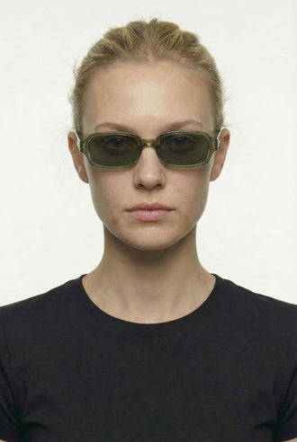 A model with blonde hair pulled back wears dark green rectangular sunglasses with a thin gold metal frame, paired with a black crewneck shirt against a plain white background.