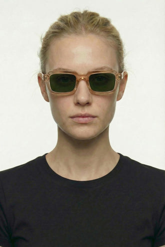 A model with blonde hair pulled back wears square sunglasses with a translucent light brown frame and dark green lenses, paired with a black crew-neck top against a plain white background.