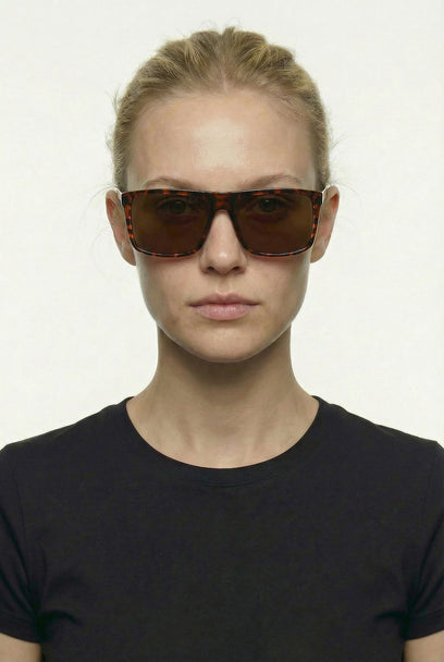 A model with blonde hair pulled back wears oversized square sunglasses with a tortoiseshell pattern and dark brown lenses, paired with a black crew-neck t-shirt against a plain white background.