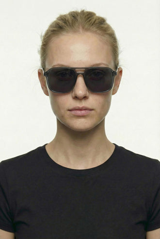 A woman with blonde hair pulled back wears dark aviator-style sunglasses with a thin metallic frame and a black crew-neck t-shirt, photographed against a plain light background.