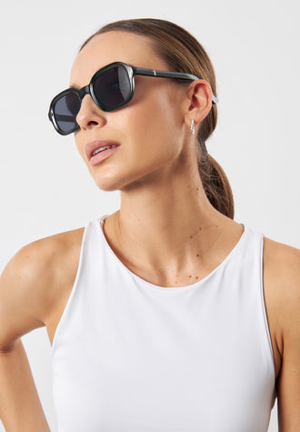 A woman wearing black cat-eye sunglasses and a white tank top, shown from the chest up against a light gray studio background.