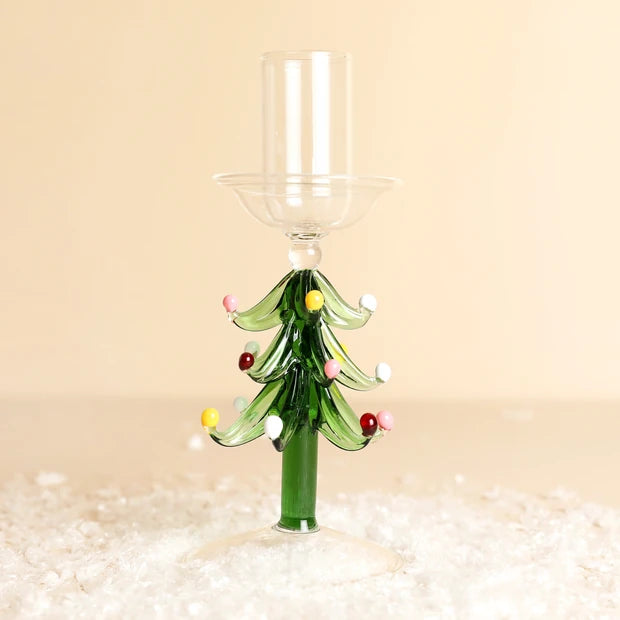 A Christmas tree-shaped glass candle holder with a clear glass candle sleeve and small colorful glass ornaments, set against a warm beige background with white faux snow at the base.