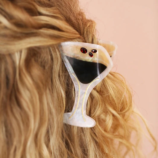 A close-up of a person's blonde hair secured with a cocktail glass-shaped hair clip. The clip is made of a translucent white and beige resin with a black center and small red gem-like accents.