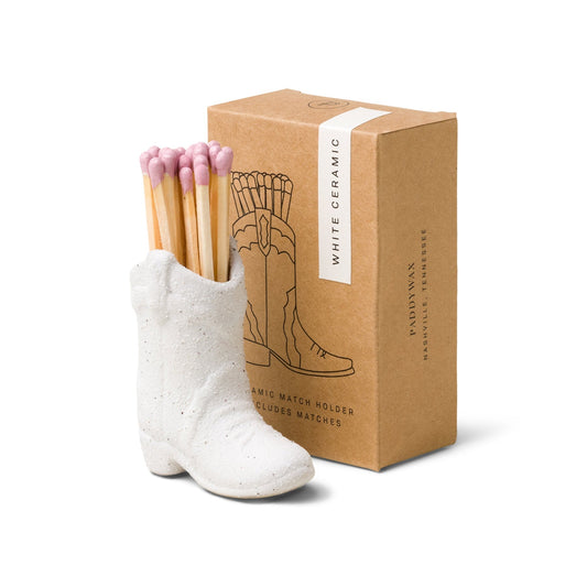 A white ceramic cowboy boot match holder with a pair of pink matches inserted at the top, placed next to a brown cardboard box with a matching design and text.