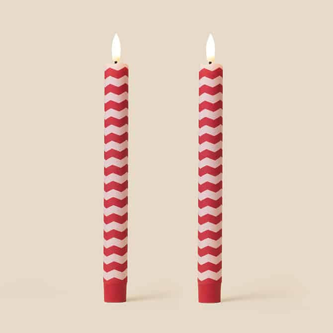 Two red and white chevron-patterned taper candles with lit flames, standing upright against a solid beige background.