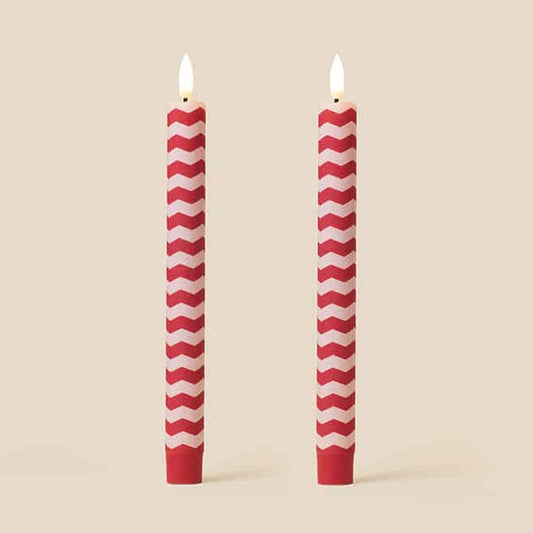 Two red and white chevron-patterned taper candles with lit flames, standing upright against a solid beige background.