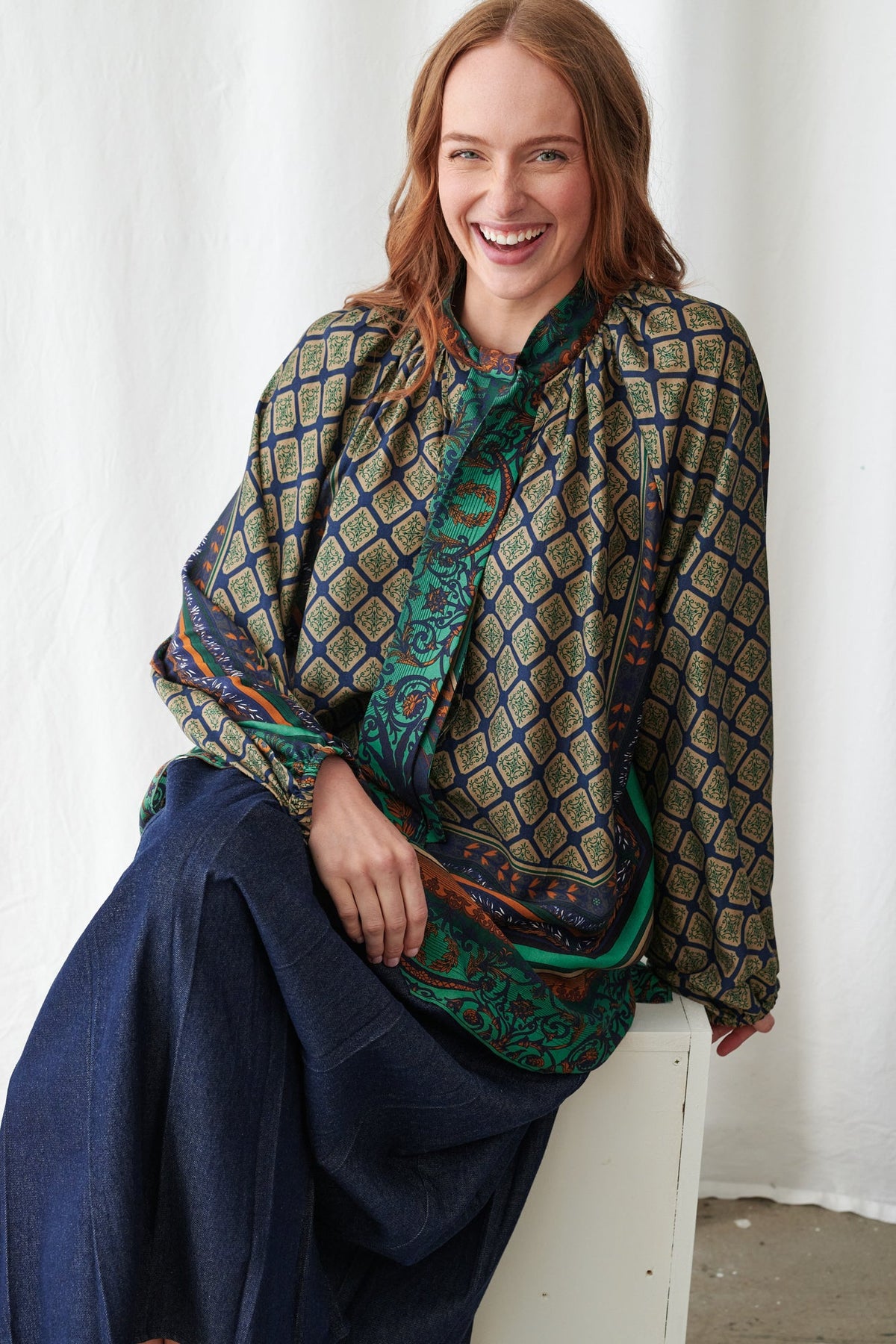 Woman wearing a patterned scarf and dark blue dress sitting on a white stool against a white curtain.