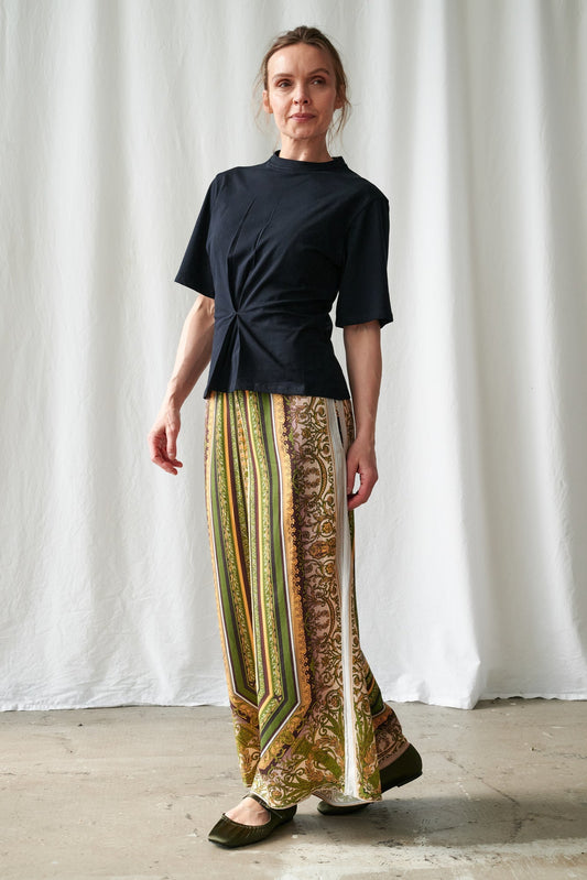 One Hundred Stars Amulet Green Palazzo Pants - Woman wearing a black top and patterned pants standing against a white curtain.