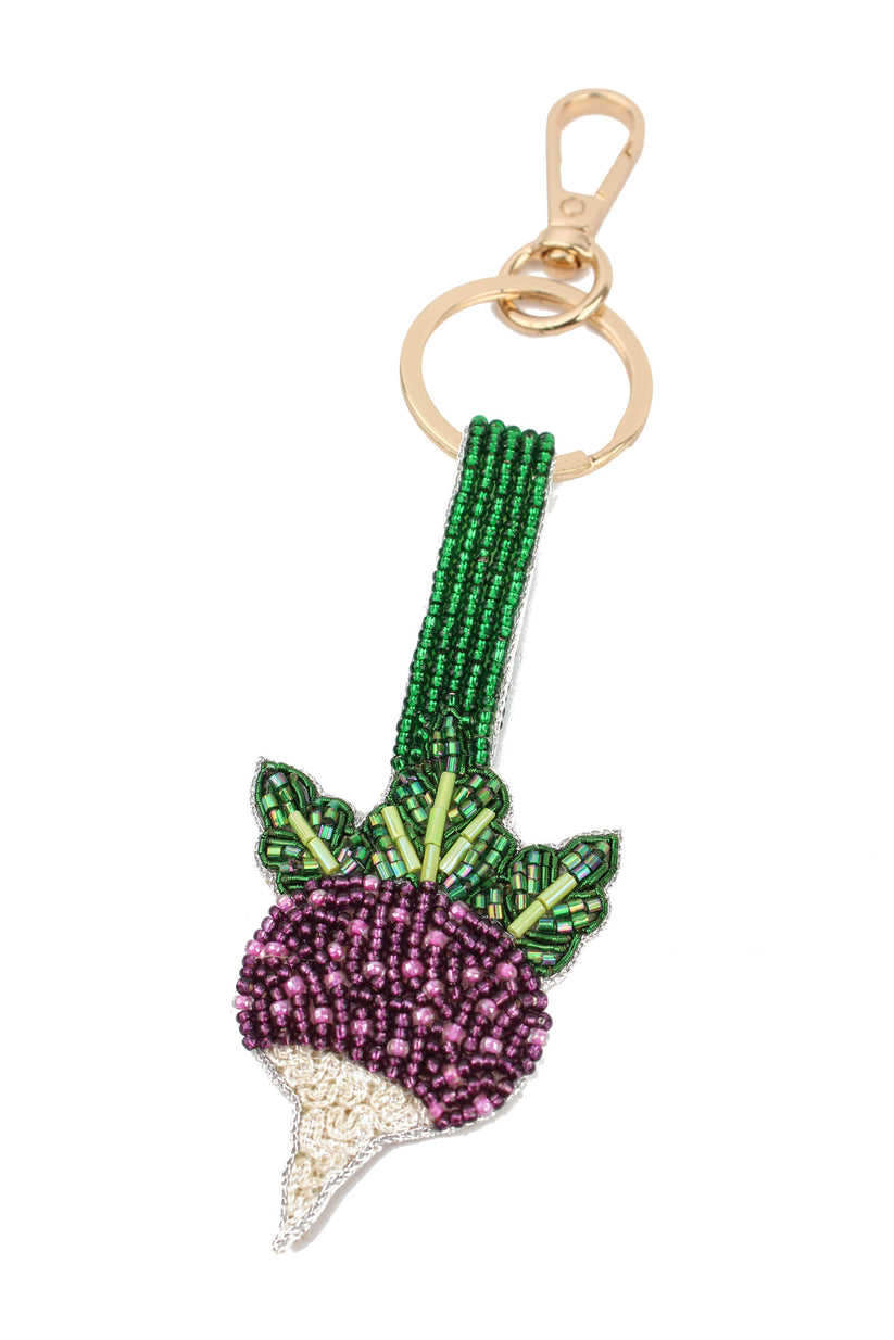 A beaded keychain charm shaped like a turnip, featuring a green seed stem, green and light green leaves, a purple beaded body, and a white beaded tip, attached to a gold-tone metal ring and clasp on a plain white background.