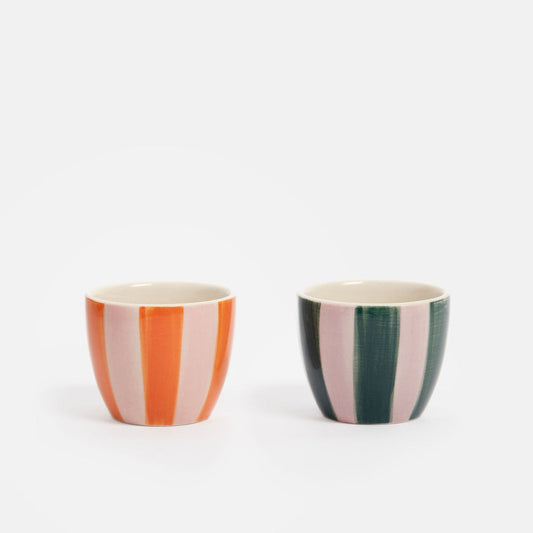 Two small ceramic cups with vertical stripes, one in orange and light pink, and the other in dark green and light pink, set against a plain white background.