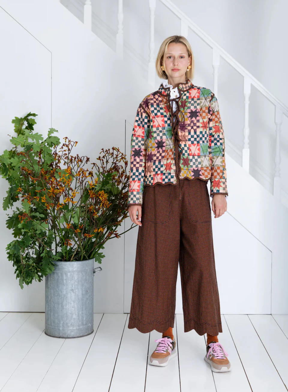 A person wearing a Bonte London Mikka Reversible Jacket in Ink & Sand and brown pants stands in a room with white walls and a plant.