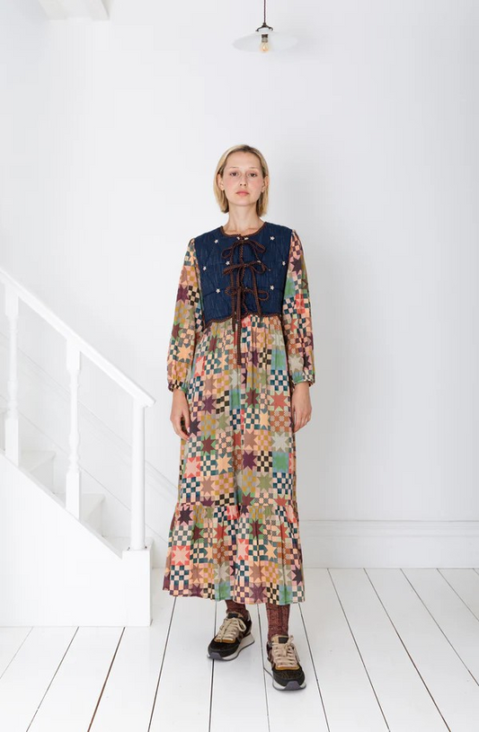 Person wearing a colorful patchwork dress with a patterned vest in a minimal white room.