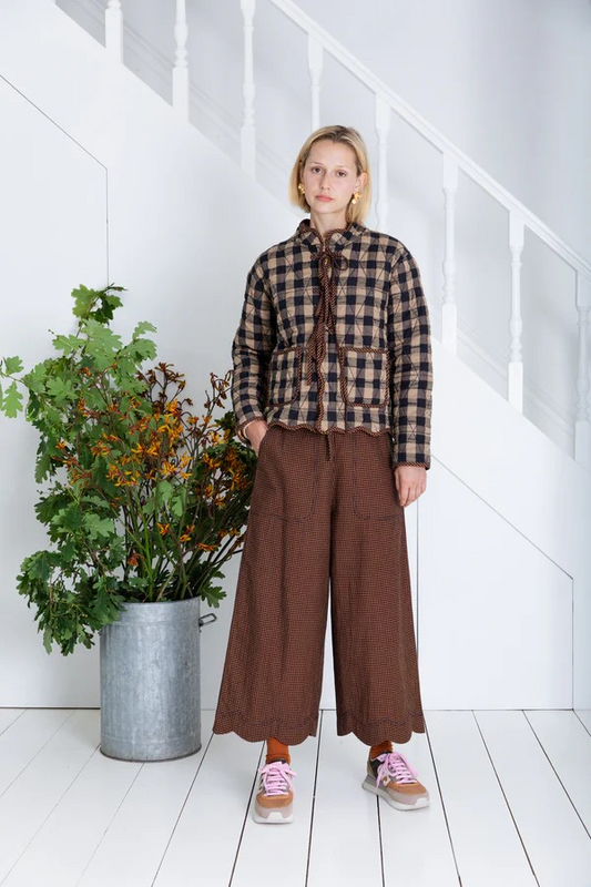 Person wearing a plaid shirt and brown pants standing next to a potted plant in a white room.