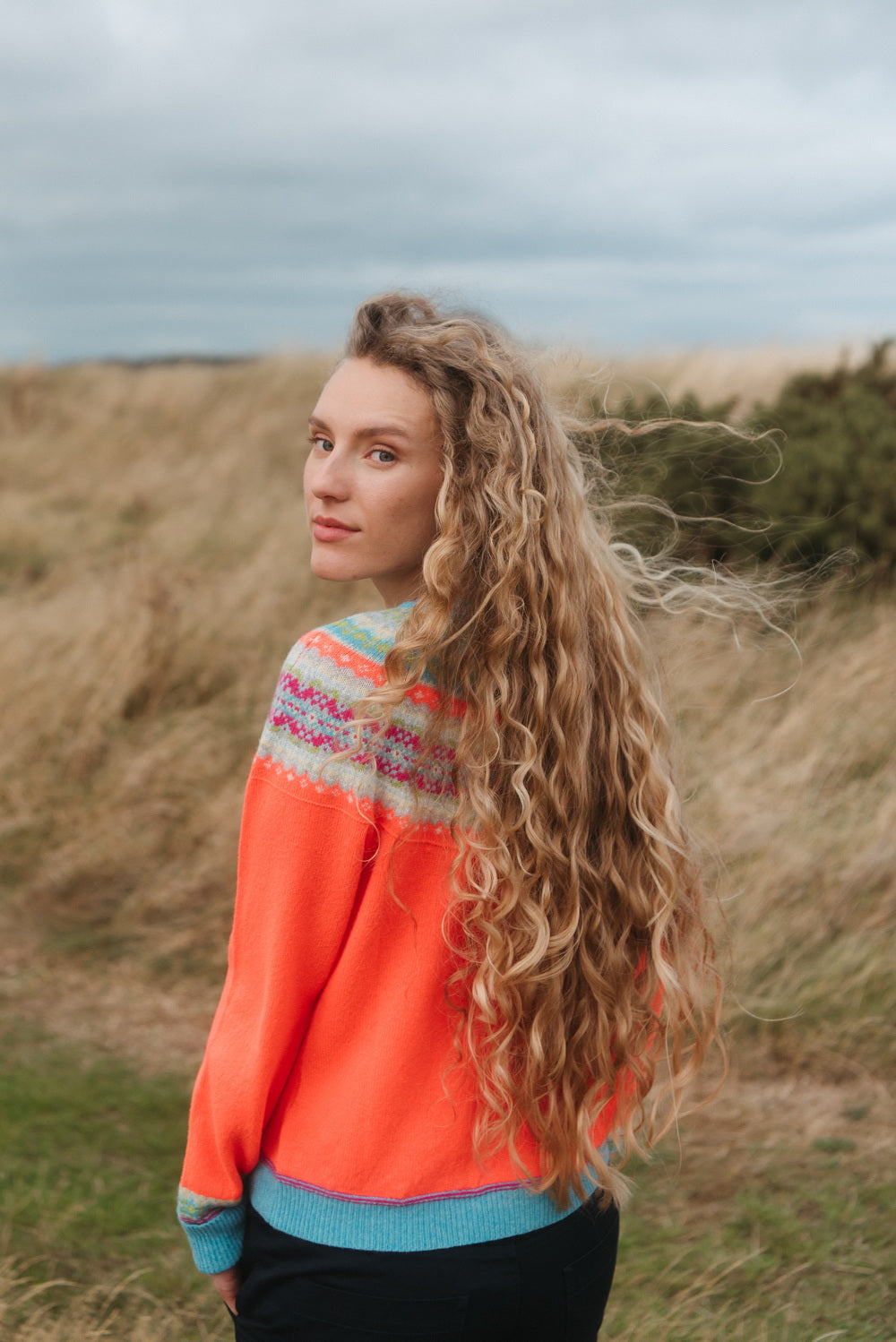 A woman with long, curly blonde hair stands outdoors in a grassy field under an overcast sky, wearing a bright orange knit sweater with a multicolor Fair Isle yoke and light blue ribbed trim.