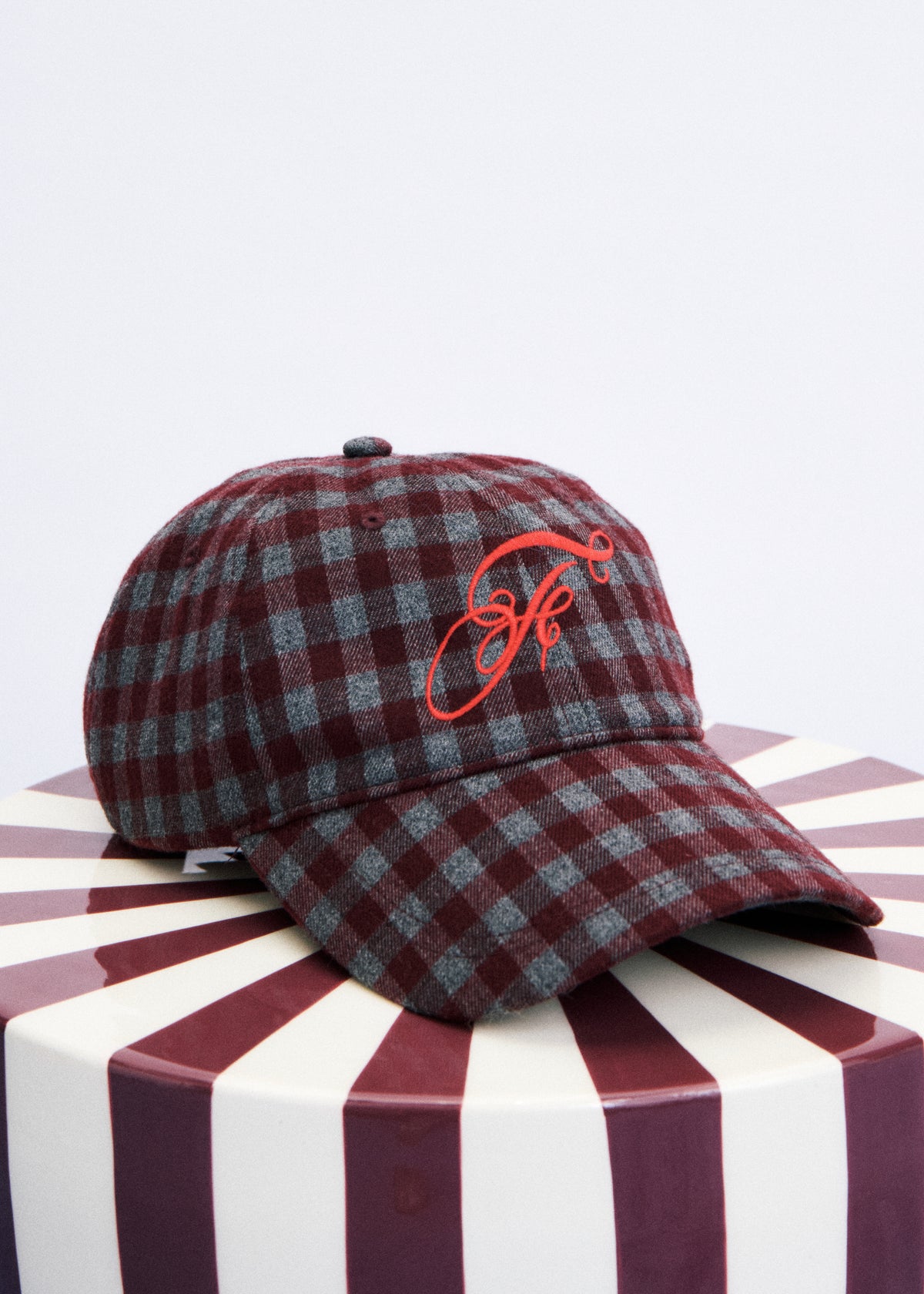 A plaid baseball cap in maroon and gray with a red embroidered monogram logo on the front, displayed on a striped round pedestal against a light gray background.