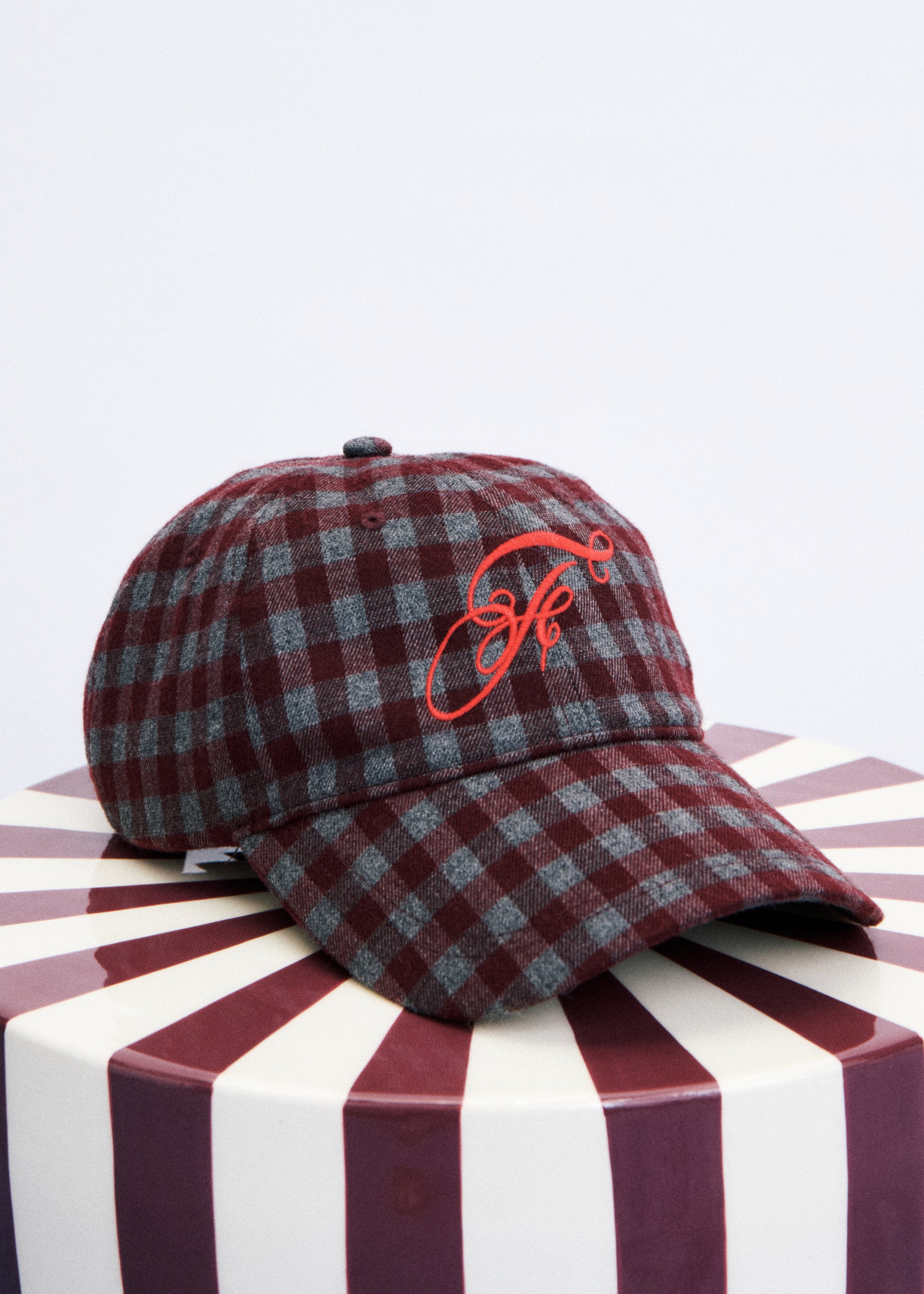 A plaid baseball cap in maroon and gray with a red embroidered monogram logo on the front, displayed on a striped round pedestal against a light gray background.