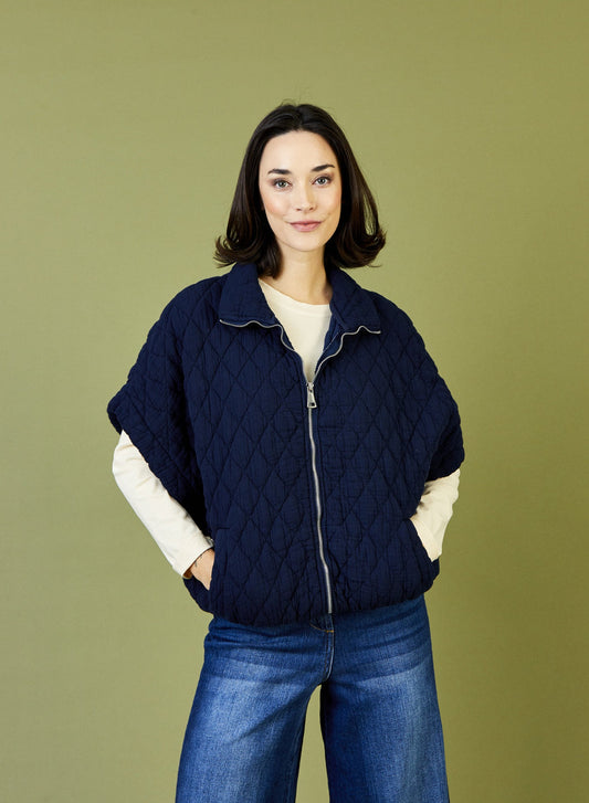 A woman modeling a navy blue diamond-quilted zip-up poncho jacket over a cream long-sleeve top and blue jeans, set against a solid olive green studio background.