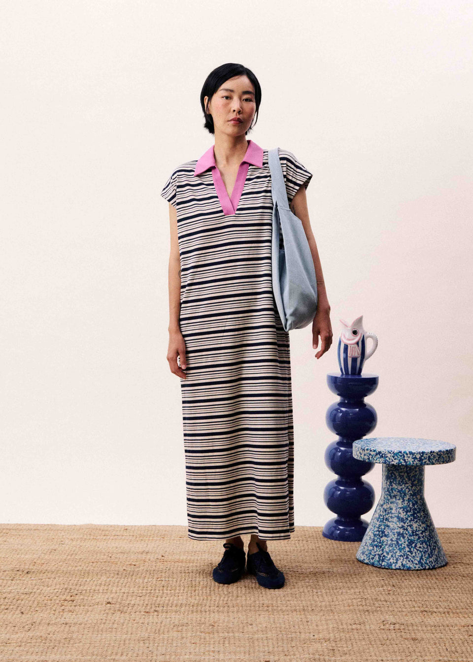 Woman in a striped dress standing next to decorative vases and a stool on a beige floor.