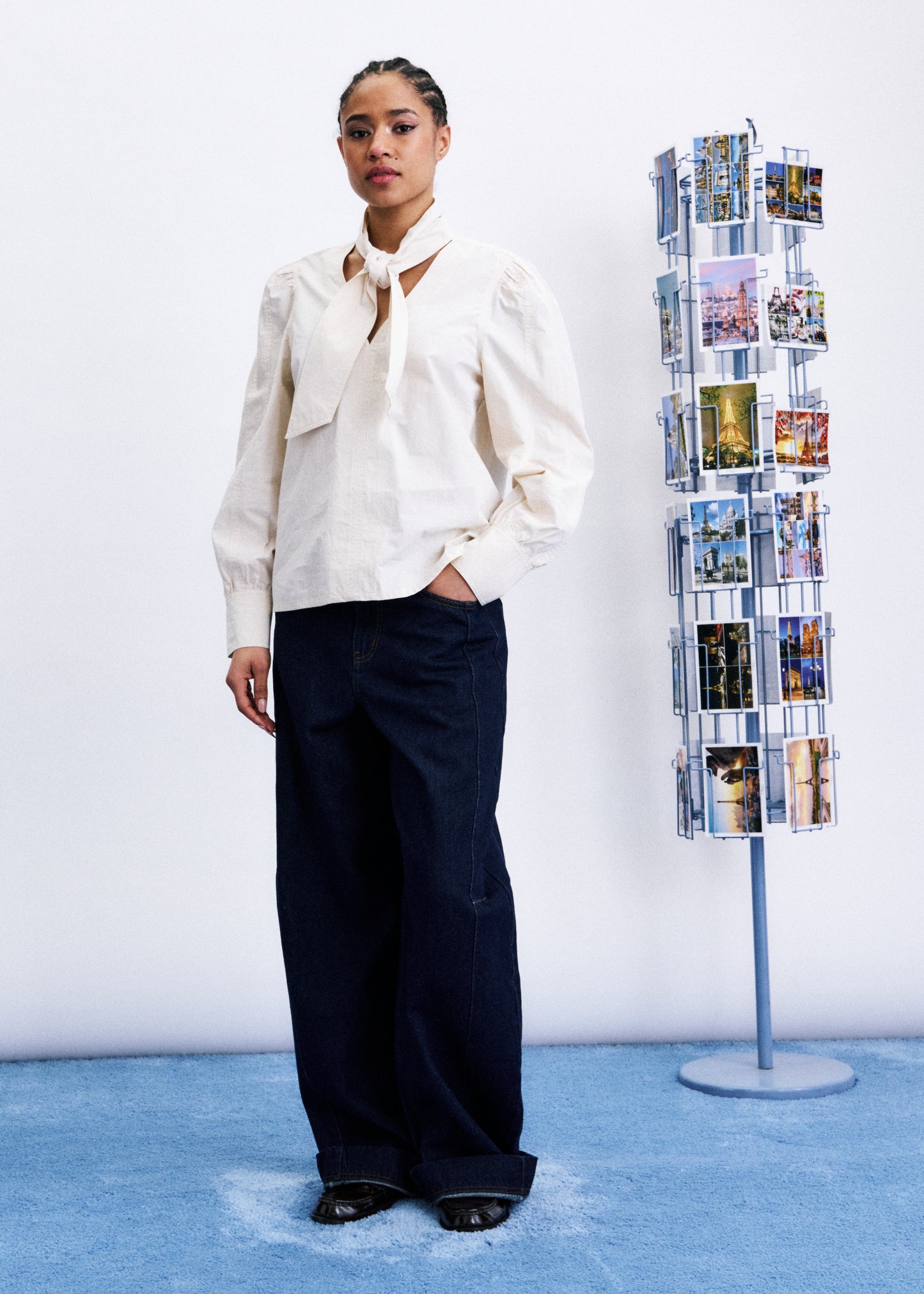 Woman in a white blouse and dark pants standing next to a magazine rack with brochures on a blue floor.