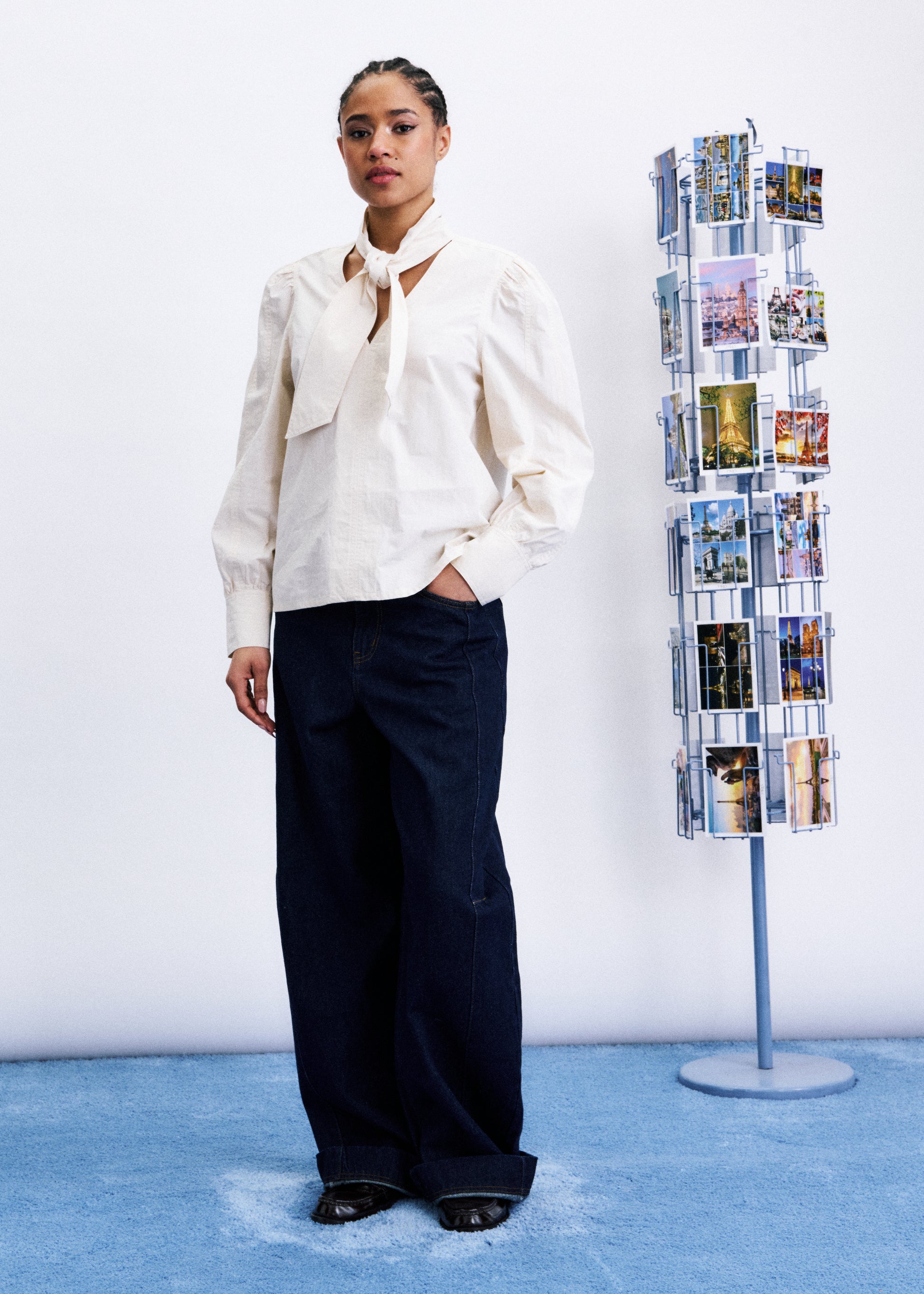 Woman in a white blouse and dark pants standing next to a magazine rack with brochures on a blue floor.