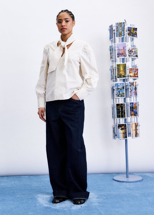 Woman in a white blouse and dark pants standing next to a magazine rack with brochures on a blue floor.