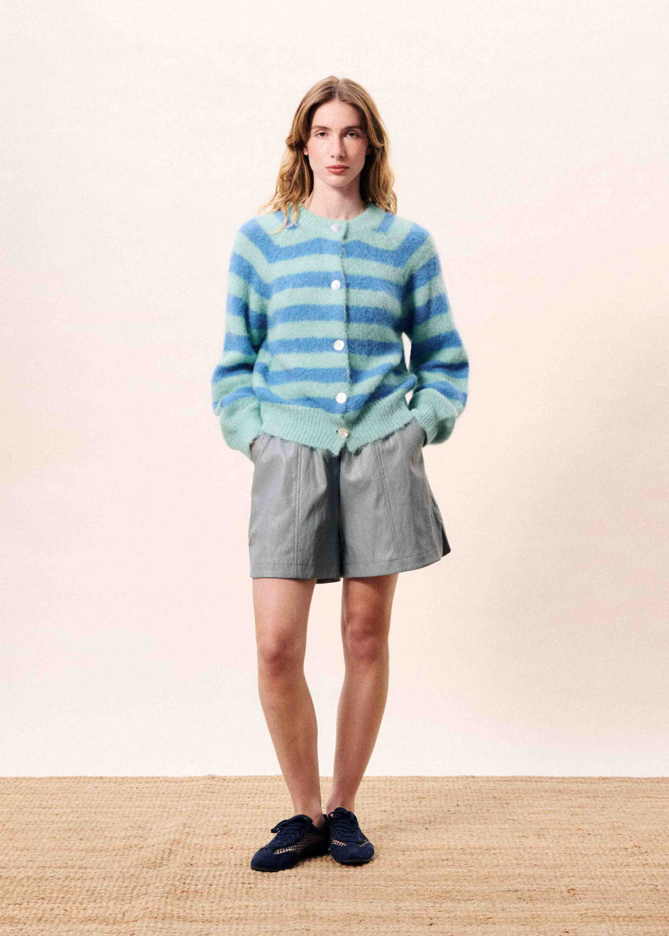 Woman wearing a blue and green striped sweater with gray shorts on a plain background