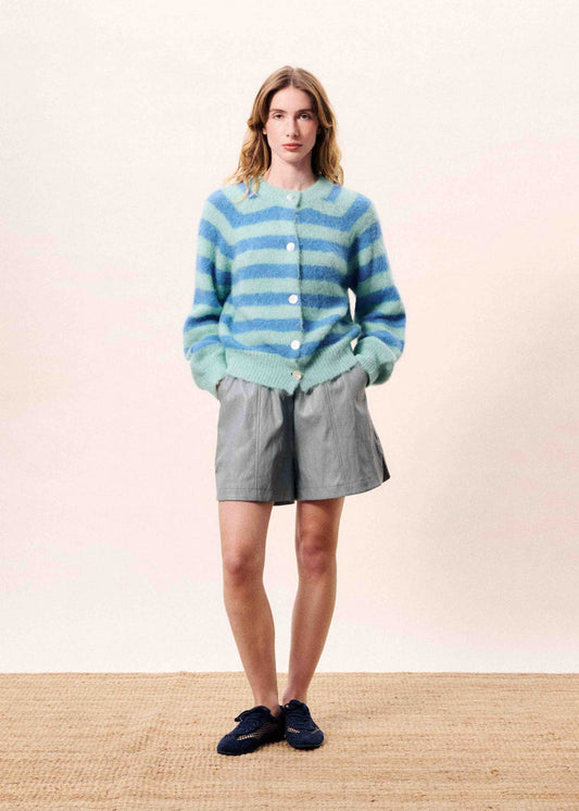 Woman wearing a blue and green striped sweater with gray shorts on a plain background